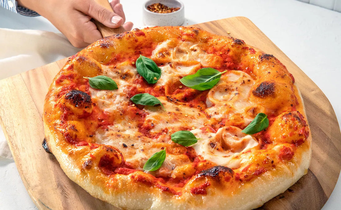 A freshly baked pizza topped with mozzarella cheese and fresh basil leaves, placed on a wooden serving board, with a hand reaching for it.